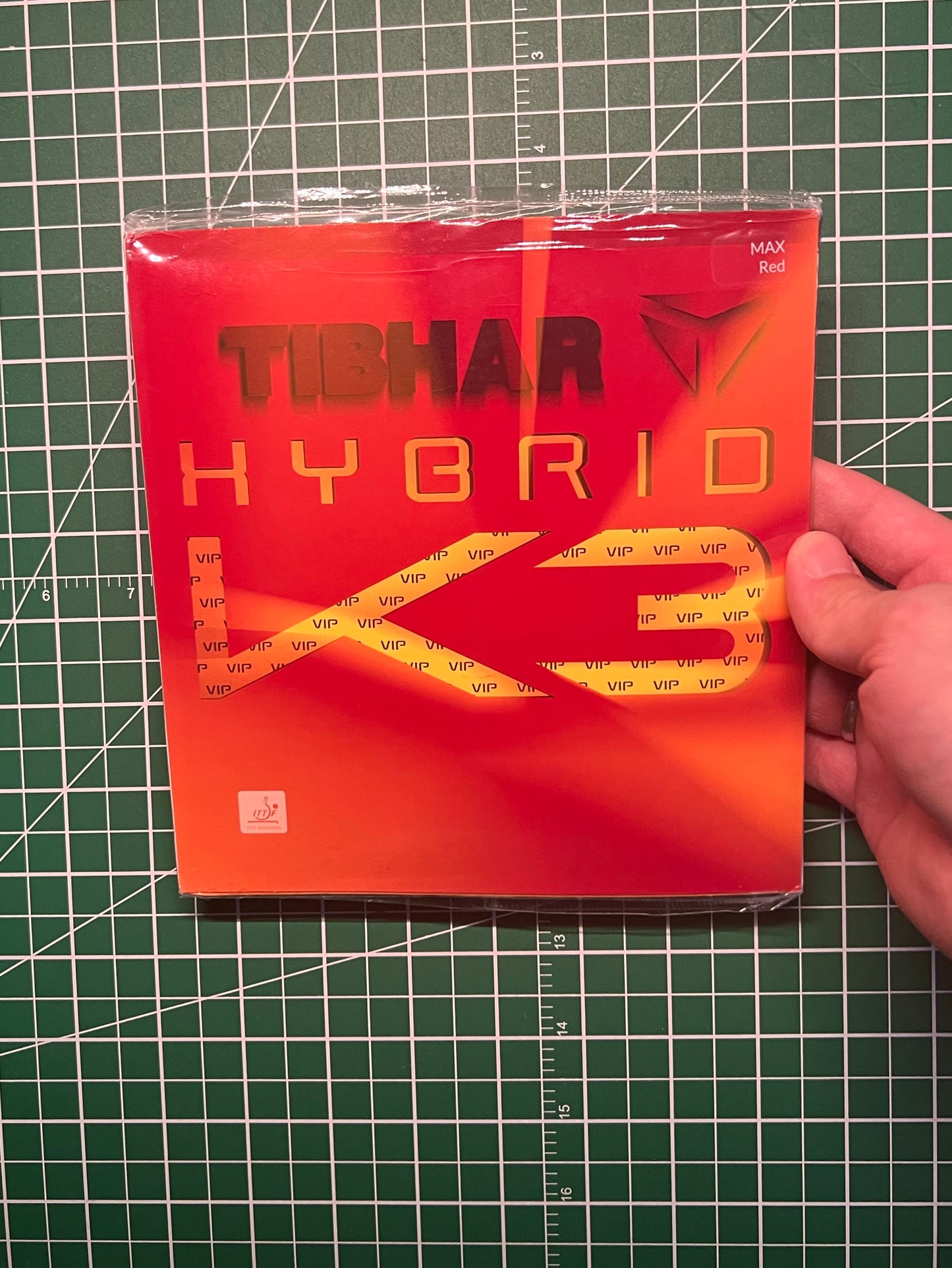 A hand holding a sealed Tibhar Hybrid K3 vip table tennis rubber against a green cutting mat background.