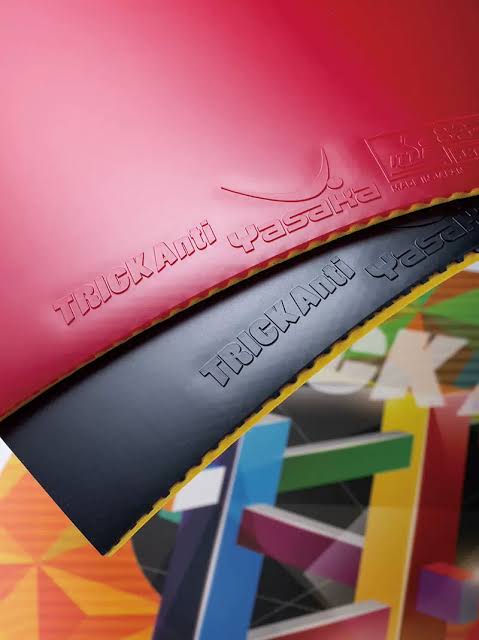 Close-up of a table tennis rubber showing two layers: a red top layer with embossed 'TRICKAnti Yasachi' branding and a black bottom layer with a yellow edge, set against a colorful geometric background.